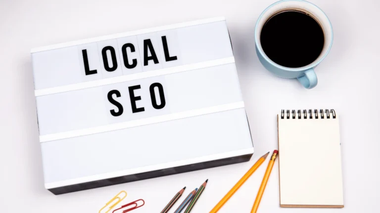 A flat-lay desk setup featuring a light-box sign reading "LOCAL SEO" beside a cup of black coffee, pencils, paper clips, and a blank spiral notepad.
